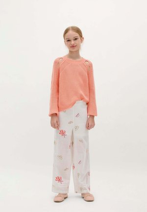 Koraalgekleurde gebreide trui met raglanmouwen, voorzien van uitsparingen. Gecombineerd met witte, wijde broek versierd met kleurrijke patronen.