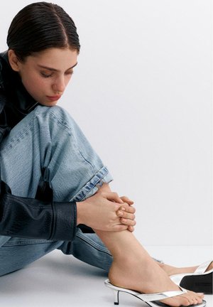 Mladá osoba sedí na zemi, má na sobě světle modré džíny a černou bundu, drží si kotník s bosýma nohama v stříbrných sandálech na nízkém podpatku na bílém pozadí.