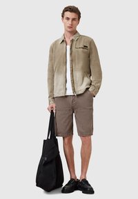 Beeži tooniga jakk esiluku ja valge T-särgiga, pruunid taskutega lühikesed püksid ning must kott; kantud mustade jalanõudega.