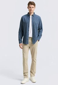 Chemise bleue à manches longues boutonnée avec une poche, associée à un pantalon beige et des baskets blanches. Texture de tissu lisse et coupe décontractée.