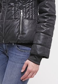 Schwarze gepolsterte Jacke mit strukturierter Oberfläche, ausgestattet mit einem Reißverschluss vorne und elastischen Bündchen. Getragen über Jeans.