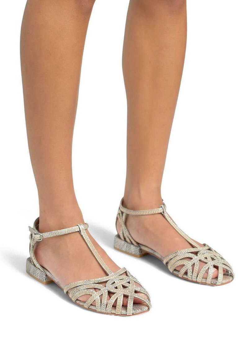 Sandalias planas con tiras en plateado metálico, con recortes decorativos, correa de tobillo ajustable, superficie texturizada y tacón bajo tipo bloque.