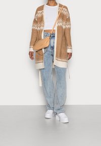 Brun stickad cardigan med vita detaljer, tillsammans med en vit croptop, ljusblå jeans och vita plattformssneakers. Beige axelväska.