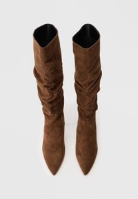 Par de botas marrones de ante hasta la rodilla con puntera afilada y diseño de caña holgada, vistas desde arriba sobre un fondo blanco.