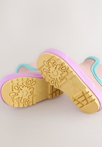 Kinderregenlaarzen met gele zolen met smiley-vlekpatronen, roze middenzolen en aquablauwe bies op een beige achtergrond.