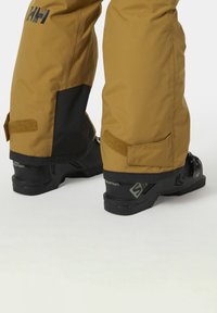 Botas de esquí negras llevadas con pantalones de nieve marrones, con una correa de Velcro y patrones texturizados. Las botas tienen marcas de la marca y un diseño resistente.