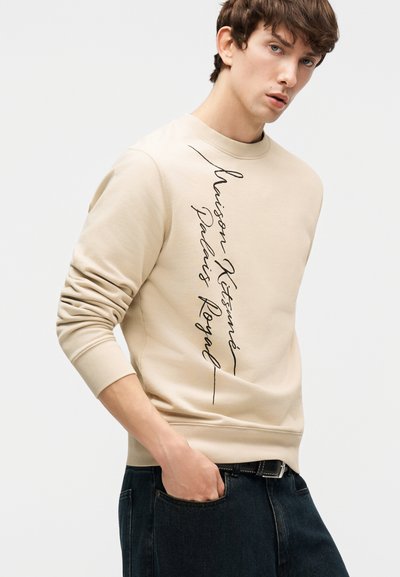 Νεαρός άνδρας με μπεζ φούτερ με μαύρο κάθετο κείμενο "Maison Kitsuné Palais Royal" και σκούρο τζιν, με το χέρι στην τσέπη, κοιτάζει πλάγια.