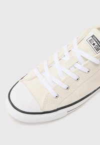 Beige suede sneaker with white laces, rounded white rubber toe cap, black trim along the sole, and a logo patch on the tongue.