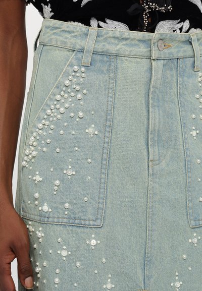 Ανοιχτό μπλε τζιν φούστα με μεγάλες μπροστινές τσέπες, διακοσμημένη με σύνολα λευκών περλών και κουμπιών σε διάφορα μεγέθη.