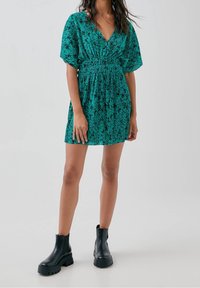Robe à imprimé floral vert avec un décolleté en V, taille cintrée et manches courtes ; associée à des bottines à talons épais noires.