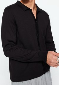 Svart långärmad polo med knäppt fram, mjukt material och platt krage. Bärs med grå byxor och synliga ringar på händerna.