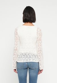 Λευκό πουλόβερ από δαντέλα με μακριά μανίκια, που διαθέτει γεωμετρικό μοτίβο και εφαρμοστή γραμμή, συνδυασμένο με μπλε τζιν παντελόνι.