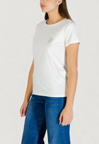 Biała bawełniana koszulka z okrągłym dekoltem i krótkimi rękawami, z ozdobnym srebrnym emblematem na lewej piersi. W połączeniu z niebieskimi dżinsami.