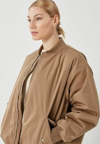 Chaqueta bomber marrón con un acabado liso y brillante, que presenta un cierre de cremallera en la parte delantera, cuello acanalado y bolsillos laterales. Corte holgado con mangas anchas.