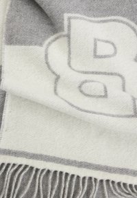 Couverture en mélange de laine mettant en avant un schéma de couleurs gris et blanc, avec des bords frangés et un grand design de logo gris sur fond blanc.