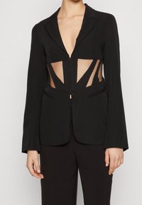 Schwarzes Blazer mit einem tiefen V-Ausschnitt, auffälligen Ausschnitten und durchsichtigen Netzabschnitten. Taillierte Passform mit strukturierten Schultern und Klappen.