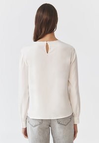Blusa beige chiara con maniche lunghe, dettaglio a chiave sul retro e texture liscia e morbida. Vestibilità sartoriale con polsini abbottonati.