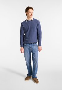 Marineblauer, gerippter Pullover mit Rundhalsausschnitt, getragen über einem hellen Hemd mit Kragen, kombiniert mit hellblauen Jeans und beigen Slippern.