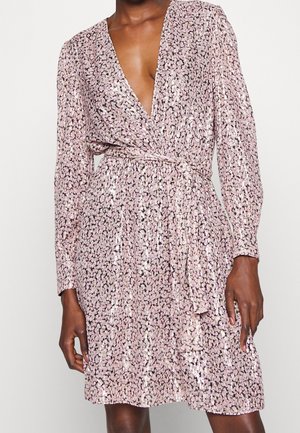 Personne portant une robe portefeuille à sequins rose clair à manches longues, avec un profond décolleté en V et une taille nouée, debout devant un fond uni.