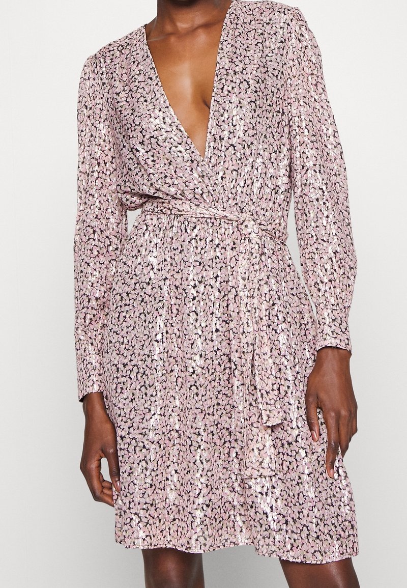 Personne portant une robe portefeuille à sequins rose clair à manches longues, avec un profond décolleté en V et une taille nouée, debout devant un fond uni.