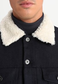 Czarna wełniana kurtka z kremowym kołnierzem z owczej skórki, z metalowymi zatrzaskami i kieszeniami na piersi, prezentująca teksturowany materiał oraz dopasowany krój.
