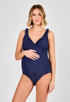 Femme enceinte portant un maillot de bain une pièce bleu marine à volants avec de petits pois noirs, posant avec une main sur son ventre sur un fond uni.