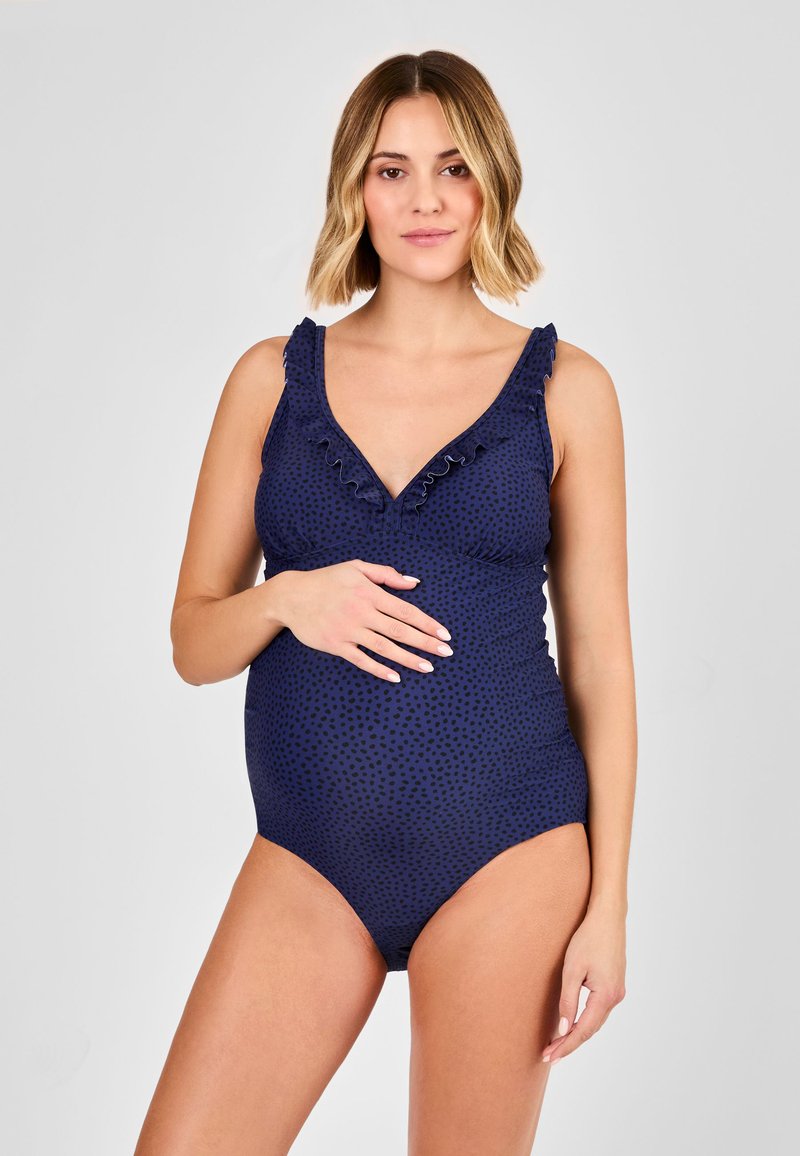 Gravid kvinde i en navyblå, flæset badedragt med små sorte prikker, poserer med den ene hånd på maven mod en ensfarvet baggrund.