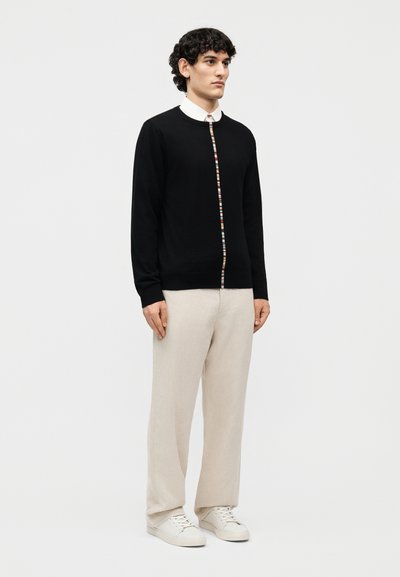 Άνδρας φοράει μαύρη ζακέτα με πολύχρωμες ρίγες μπροστά, λευκό πουκάμισο με γιακά, μπεζ παντελόνι και λευκά αθλητικά παπούτσια, σε γκρι απλό φόντο.