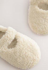 Chaussons crémeux et duveteux en matière polaire douce et texturée, avec des découpes sur les côtés pour la respirabilité et un port facile.