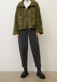 Grøn fleeceforet jakke med kviltede paneler, to frontlommer og høj krave. Bar over sorte poserede jeans og kraftige sorte støvler.