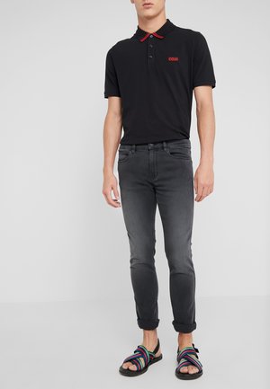 Uomo con polo nera con rifiniture rosse, jeans grigio scuro arrotolati e sandali a righe multicolore in piedi davanti a uno sfondo semplice.