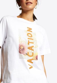Witte katoenen t-shirt met een bloemenprint en "VACATION" in grote oranje letters en heldere rhinestone accenten. Korte mouwen en ronde halslijn.