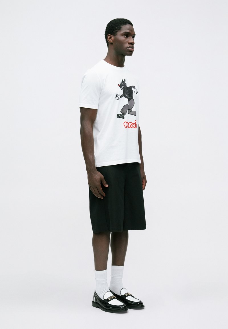 Witte katoenen t-shirt met een zwart cartoonafbeelding, gecombineerd met zwarte knielange shorts en zwart-witte loafers. Witte sokken zichtbaar.