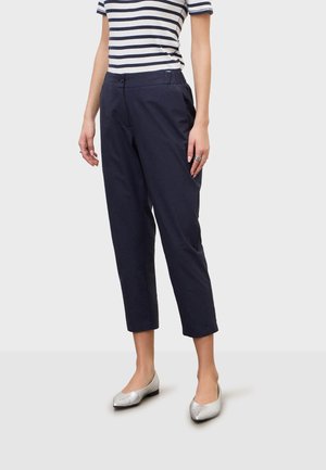 Osoba oblečená v námornícky modrých skracovaných nohaviciach, bielom a námornícky modrom pruhovanom tričku s krátkym rukávom a striebornej bezpej obuvi stojaca na jednofarebnom pozadí.