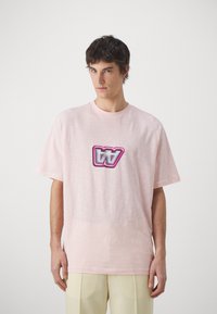 Camiseta de manga corta rosa claro en un corte relajado, con un logotipo gráfico central en tonos rosa y gris. Textura suave con un acabado liso.