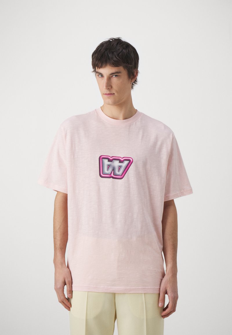 Camiseta de manga corta rosa claro en un corte relajado, con un logotipo gráfico central en tonos rosa y gris. Textura suave con un acabado liso.