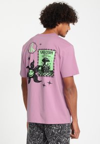 Paarse T-shirt met korte mouwen met een groot zwart grafisch ontwerp op de achterkant, inclusief een ingelijste illustratie en gestileerde elementen.