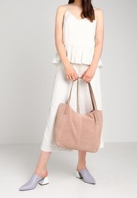 Sac cabas en suède rose avec une texture douce, des anses en cuir marron et un design spacieux. Le modèle porte un haut plissé crème et un pantalon à jambes larges.