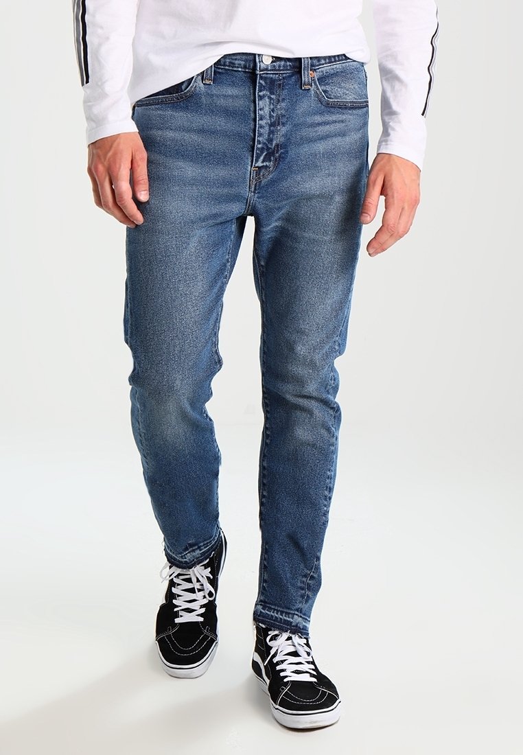 Blue denim jeans with a straight fit, faded texture, five-pocket design, and subtle stitching details, paired with black sneakers.