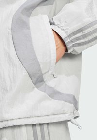 White athletic jacket featuring a textured material, pocket detail with a contrast gray accent, and elastic cuffs with a drawstring at the hem.