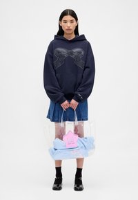 Granatowa bluza z kapturem o luźnym kroju z naszytym wzorem wstążki. Modelka ma na sobie dżinsową spódnicę i trzyma przezroczystą torbę typu tote z różowymi akcentami oraz jasnoniebieskie ubrania.