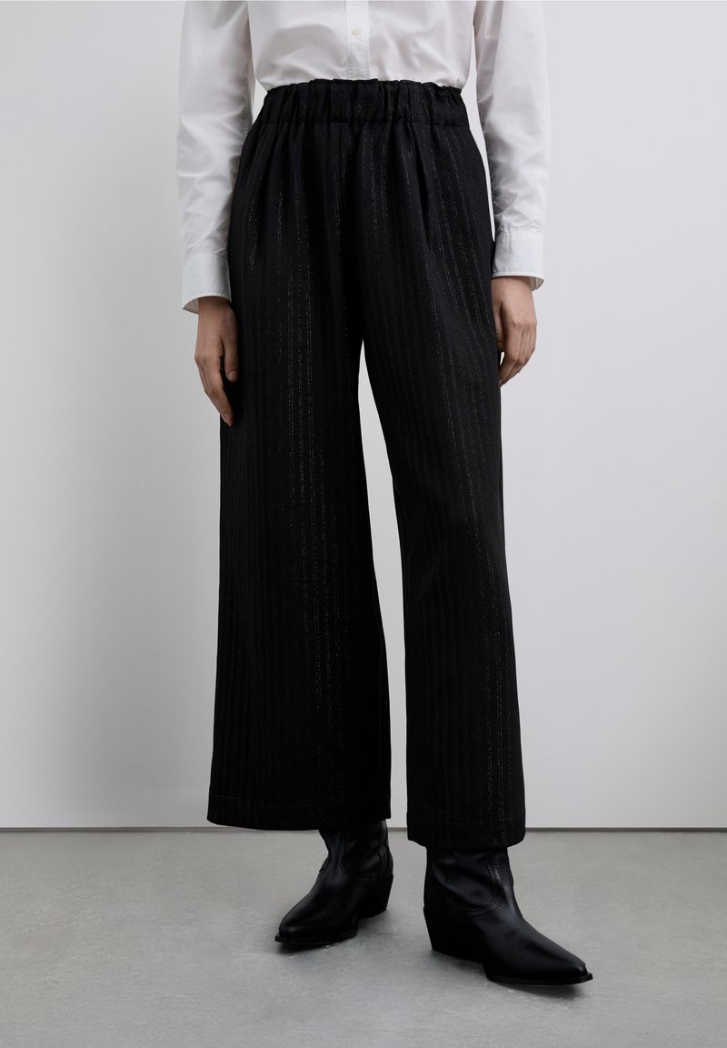 Pantalones anchos negros con cintura plisada. Tejido texturizado con líneas verticales. Combinados con botines negros ajustados.