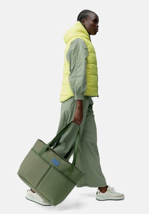Grønn kanvas totebag med to frontlommer, blå etikett og lange stropper, båret av en person iført en gul puffervest og grønt antrekk.