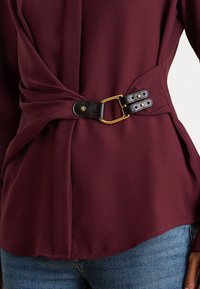 Vīna krāsas blūze no gludas auduma, ar melnu ādas jostu ar zelta sprādzi, drapētu dizainu un savāktām sānu detaļām.