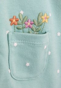 Helles blaues Material mit einer Tasche, die bestickte Blumen in Orange, Pink, Gelb und Grün zeigt, sowie kleinen weißen Blumenmotiven.
