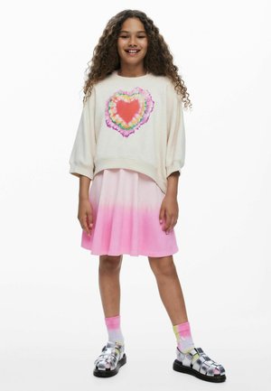 Hosszú, göndör hajú lány, krémszínű pulóvert visel, melyen színes szív látható, rózsaszín ombre szoknyát, batikolt zoknit és ezüstszínű szandált, áll és mosolyog.