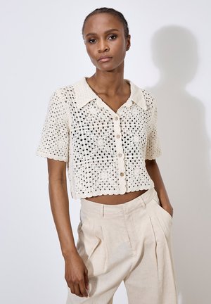 Beige gehäkelte Strickjacke mit Kragen, kurzen Ärmeln und Knopfverschluss. Das Design hat ein florales Muster und gewellte Kanten.