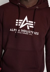 Burgunder hettegenser med hvit logo "ALPHA INDUSTRIES." Har justerbare snorer og en frontlomme, laget av myk stoff.