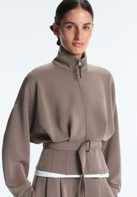 Donna con capelli scuri legati all'indietro, indossa una giacca taupe con zip e cintura a vita alta abbinata a pantaloni coordinati, di fronte a uno sfondo semplice.