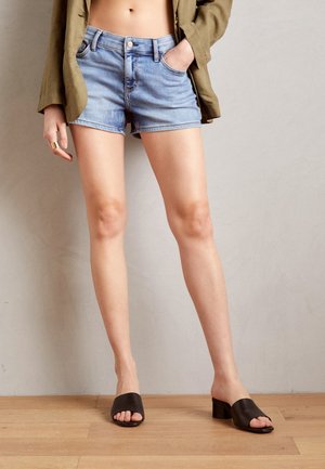Person iført lyseblå cowboyshorts, olivengrøn jakke, sorte åbne tå-hælsandaler, stående mod en enkelt væg på trægulv.